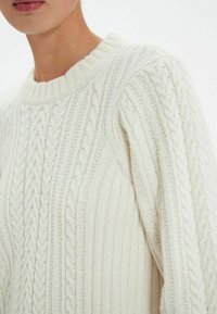 Pull en maille torsadée de couleur crème, avec une texture côtelée, un col rond et des motifs de tressage intriqués. Tissu épais, design confortable.
