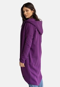 Fioletowa, długorękawowa bluza z kapturem o luźnym kroju. Posiada dyskretne boczne zamki i strukturalną powierzchnię. Nosi się na dżinsach.