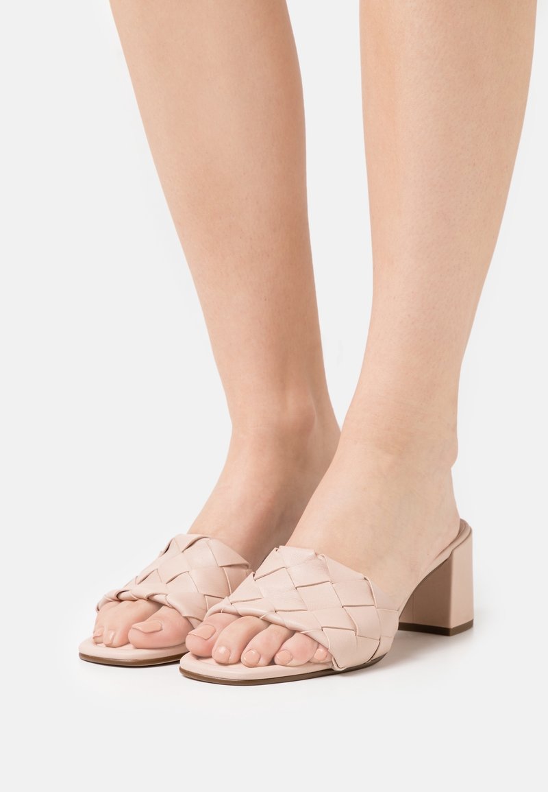 Pieds portant des sandales tissées rose clair à talons blocs et à bouts ouverts sur fond blanc.