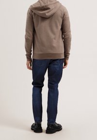 Man i brun hoodie, mörkblå jeans och svarta skor som står vänd bort mot en enfärgad bakgrund.