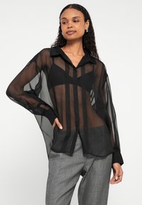 Blusa preta transparente com botões, mangas compridas, gola e padrão de riscas verticais. Usada por cima de um top preto e calças cinzentas com padrão.
