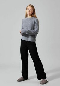 Grau gestrickter Pullover mit gerippter Textur, rundem Halsausschnitt und langen Ärmeln. Kombiniert mit schwarzen, weit geschnittenen Hosen und grauen Hausschuhen.