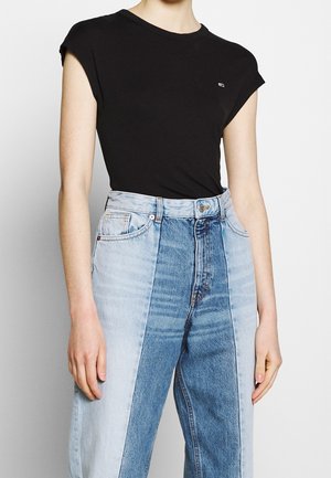 Personne portant un haut noir sans manches rentré dans un jean bleu bicolore taille haute, debout devant un fond uni.