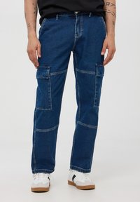 Niebieskie dżinsowe spodnie cargo z wieloma kieszeniami, prostym krojem nogawek oraz widocznymi detalami szwów. Noszone z białymi i szarymi sportowymi butami typu low-top.