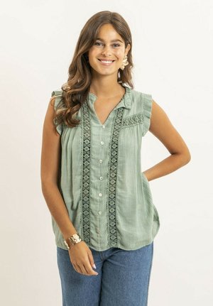 Femme souriante aux longs cheveux bruns porte un chemisier vert sans manches avec des détails en dentelle, un jean bleu, de grandes boucles d'oreilles florales et un bracelet en or.