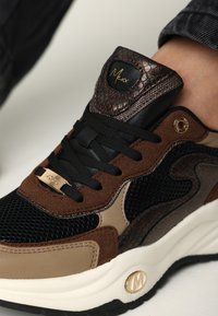 Hnědé a černé tenisky s prvky z síťoviny a semiše, lesklými akcenty a zlatou logově deskou. Bílá podrážka s texturovanými detaily.