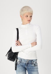 White ribbed long-sleeve top with high collar, paired with blue denim jeans. Model carries a black leather handbag with subtle detailing.