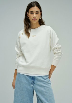 Mujer con sudadera blanca con botones dorados y logo "SALSA", combinada con jeans azul claro, de pie contra un fondo liso.