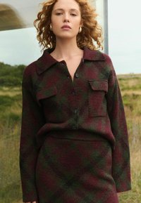 Plaid wool sweater with a collar, buttons, and two front pockets; colors include burgundy and green with a textured finish.