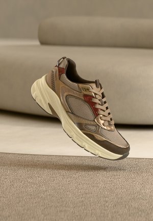 Beige og brun sportssko med røde accenter og tyk cremefarvet sål, som svæver over et tæppebelagt gulv, beige sofa i baggrunden.