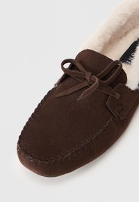 Zapatilla mocasín de ante marrón con detalles de costura en la punta, lazo atado y suave forro de lana blanca visible en el interior.