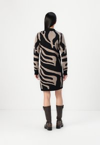 Femme aux longs cheveux noirs portant une robe à motifs abstraits noirs et beiges et des bottes montantes marron foncé, debout contre un mur blanc.