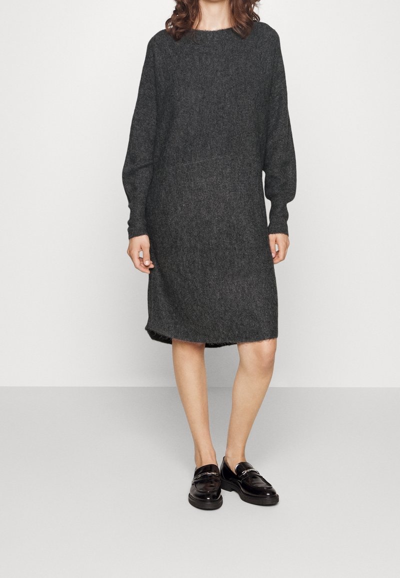 Mörkgrå stickad klänning med långa ärmar, rund halsringning och lätt fransad nederkant, i kombination med svarta loafers. Slät textur.
