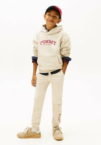 Beige Kapuzenpullover mit "TOMMY HILFIGER"-Logo, kombiniert mit passenden beigen Jogginghosen und hellbraunen Sneakers. Eine Kappe in Rot und Marineblau rundet das Outfit ab.