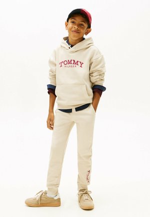 Beige hættetrøje med "TOMMY HILFIGER" logo, parret med matchende beige sweatpants og tan sneakers. Kasket med røde og marineblå farver fuldender looket.