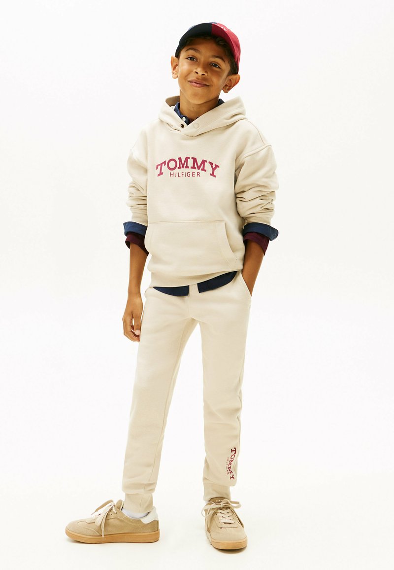 Beige Kapuzenpullover mit "TOMMY HILFIGER"-Logo, kombiniert mit passenden beigen Jogginghosen und hellbraunen Sneakers. Eine Kappe in Rot und Marineblau rundet das Outfit ab.