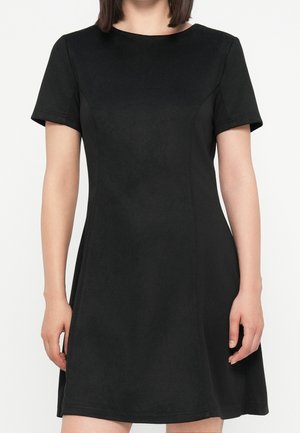 Frau trägt ein schwarzes, kurzärmeliges, knielanges Kleid mit rundem Ausschnitt vor einem schlichten hellen Hintergrund.