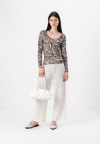 Top de manga larga con un patrón de cebra en marrón y beige, combinado con pantalones anchos blancos y semitransparentes. La modelo sostiene un bolso blanco curvado.