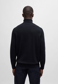 Črna pletena turtleneck pulover z rebrastim ovratnikom in manšeti, ki se ponaša z vitkim krojem. Tkanina se zdi gladka in ima mehak otip.
