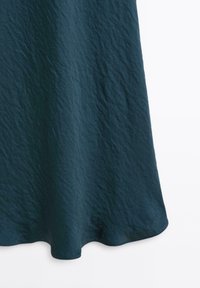 Tessuto di un verde scuro con una texture leggermente increspata, caratterizzato da un design fluido e un orlo arrotondato.