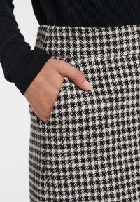 Falda de pata de gallo en blanco y negro con una mano visible en un bolsillo, que presenta una tela texturizada y un diseño ajustado en la cintura.