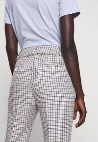 Personne portant un pantalon à carreaux taille haute avec des poches arrière boutonnées et une chemise bleu clair à manches courtes, debout devant un fond uni.