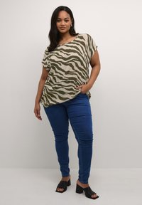 Maglietta beige svasata con strisce zebrate verdi, abbinata a jeans skinny blu e sandali neri con tacco a blocco.