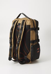 Brown fabric backpack with black mesh and red accents. Features padded straps, side pocket, and rounded design for comfort and support.
