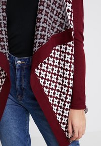 Burgundy kofta med ett mönstrat framstycke som har vita och svarta blommönster. Mjuk tyg, öppen framsidan och långa ärmar.