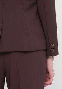 Burgundi puku, jossa on sileä pinta, kultaiset nappikiinnitykset hihansuissa ja muotoiltu malli, joka ulottuu vyötärölle.