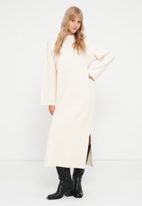 Robe longue de couleur crème avec un col montant et de larges manches. Elle comporte des fentes latérales et est accompagnée de bottes noires hauteur genou.