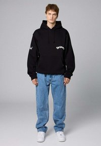 Czarna bluza z kapturem, ze sznurkiem i kieszenią z przodu, z napisem "grimey". Połączona z jasnoniebieskimi, luźnymi dżinsami i białymi sneakersami.