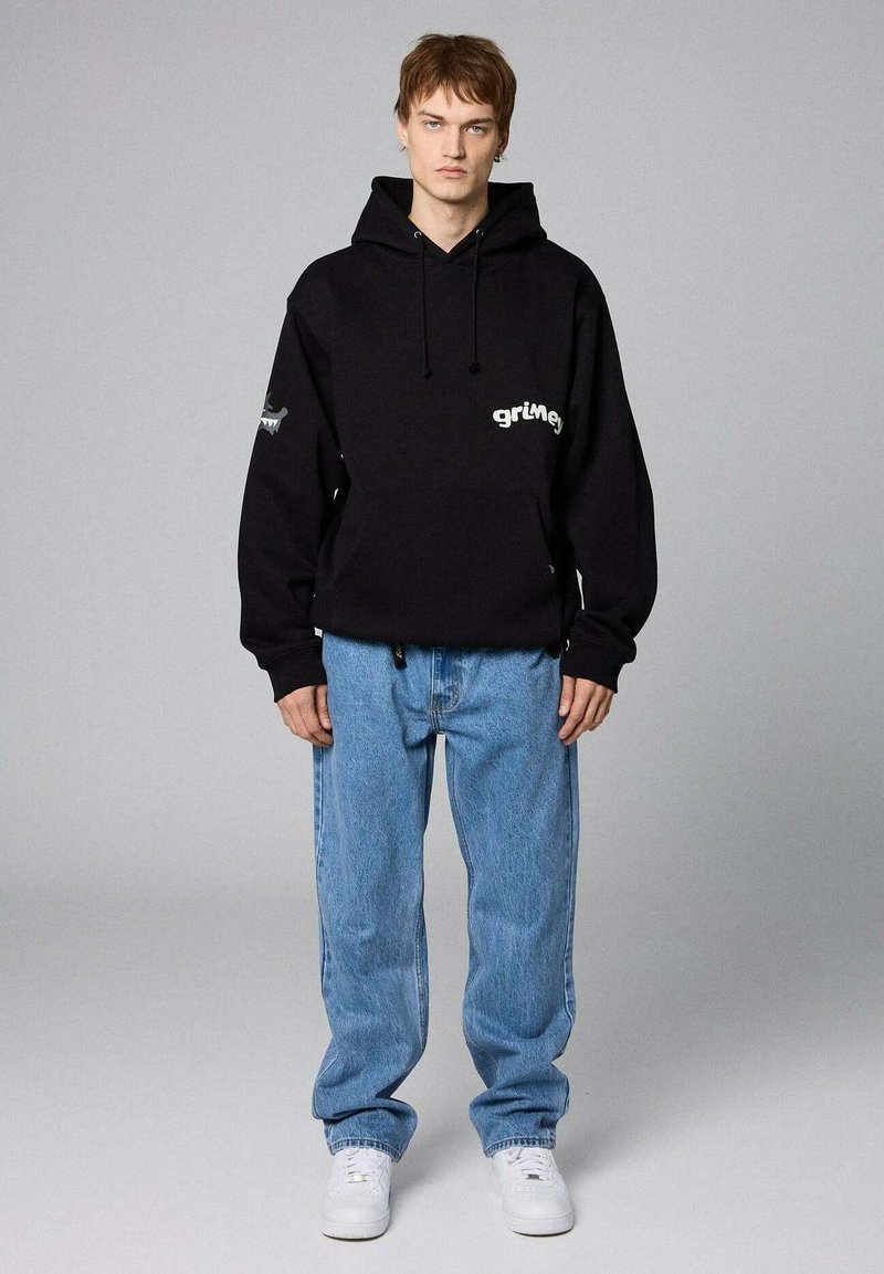 Czarna bluza z kapturem, ze sznurkiem i kieszenią z przodu, z napisem "grimey". Połączona z jasnoniebieskimi, luźnymi dżinsami i białymi sneakersami.