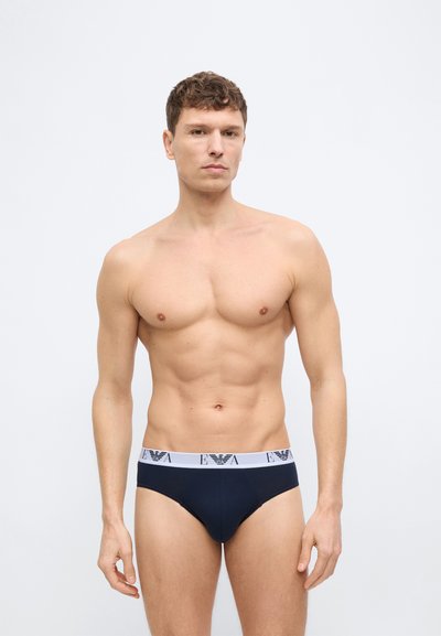 Lühikeste lokkis juustega mees, kes kannab musti bokserpükse valge bränditud vööga, seisab ühevärvilise tausta ees.