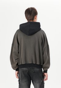 Sweatshirt com capuz, apresentando um capuz preto e um corpo verde oliva escuro, corte largo, punhos e bainha canelados. Usado com jeans cinza desgastados.