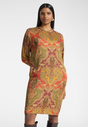 Vrouw draagt een jurk met lange mouwen tot op de knie met een paisley-patroon in rood, geel en groen, geaccentueerd met gouden oorbellen en een ketting.