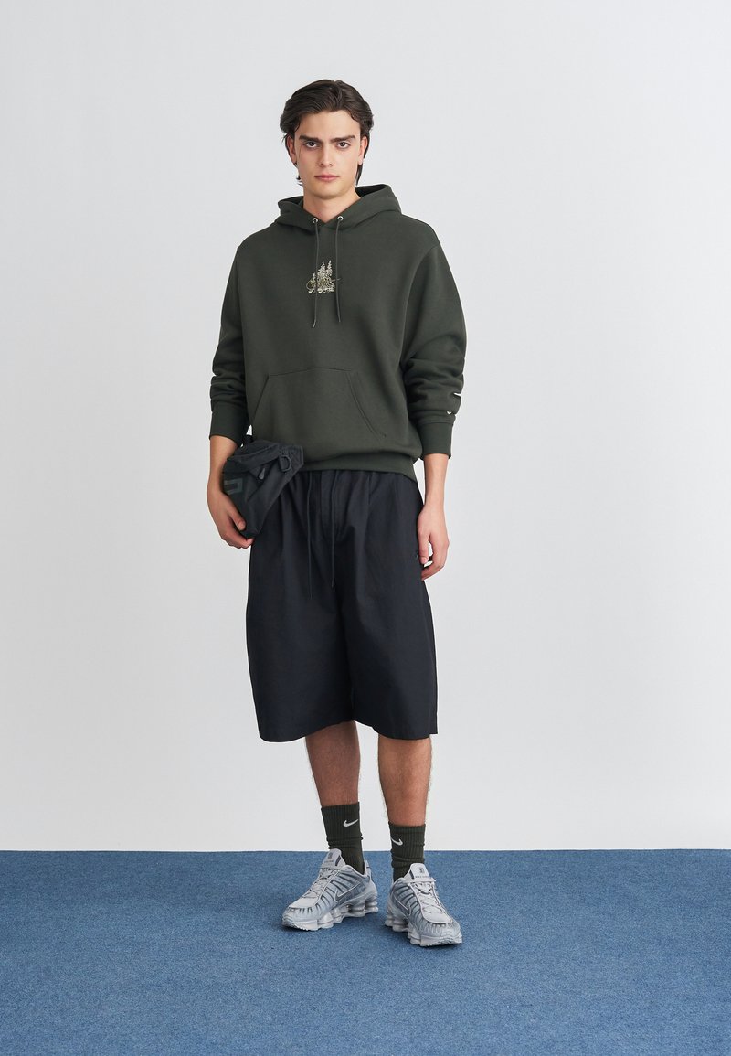 Σκούρο πράσινο φούτερ με κουκούλα και μπροστινή τσέπη, διακοσμημένο με κεντήματα, συνδυασμένο με μαύρες βερμούδες μέχρι το γόνατο και γκρι αθλητικά παπούτσια.