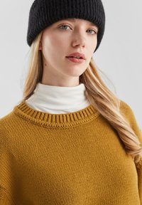 Mosterdgele gebreide trui met een geribbelde halslijn, gedragen over een witte turtleneck. Model draagt een zwarte geribbelde beanie.