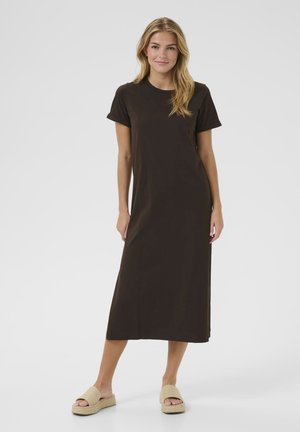 Robe brune, à manches courtes, de longueur genou en tissu doux. Dotée d'un col rond et d'une silhouette droite. Portée avec des sandales de couleur claire.