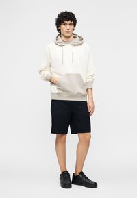 Νέος άνδρας που στέκεται, φορώντας μπεζ φούτερ με διχρωμία, μαύρο σορτς και μαύρα αθλητικά παπούτσια, με το χέρι στην τσέπη του φούτερ, μπροστά σε απλό φόντο.