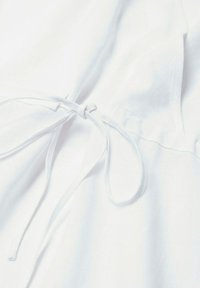 Gros plan sur un tissu de vêtement blanc avec un nœud noué fait de fines bandes de tissu au niveau de la couture centrale.