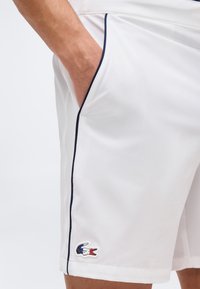 Λευκά σορτς με ναυτικό περιθώριο και μικρό κεντημένο λογότυπο κροκόδιλου κοντά στο τελείωμα, φορεμένα με ένα χέρι στην πλαϊνή τσέπη.