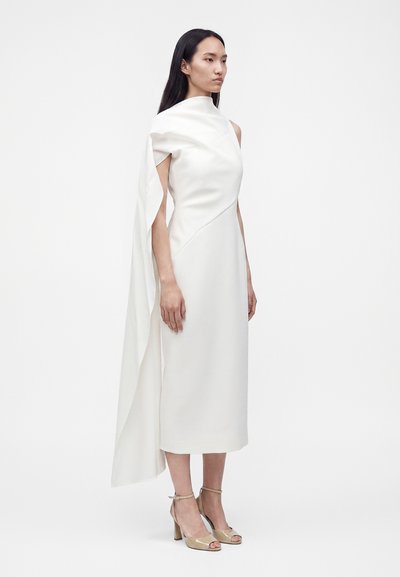 Femme en robe midi blanche avec un col montant et un long détail de cape fluide, portant des sandales à talons hauts beiges et ouvertes aux orteils.