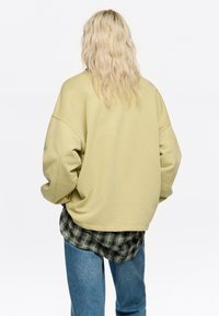 Gai zaļš pārmērīgi liels džemperis ar kritušām plecu šuvēm, teksturēta auduma, ar izliektu apakšmalu, uzvilkts pāri ruļļainai flaneļjakai un džinsiem.