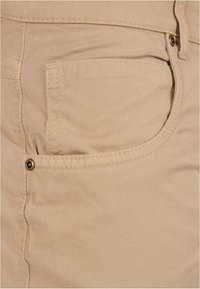 Beige puuvillakangas, jossa on näkyvät saumat ja sivutasku, metallinen napin korostus taskun aukon lähellä.