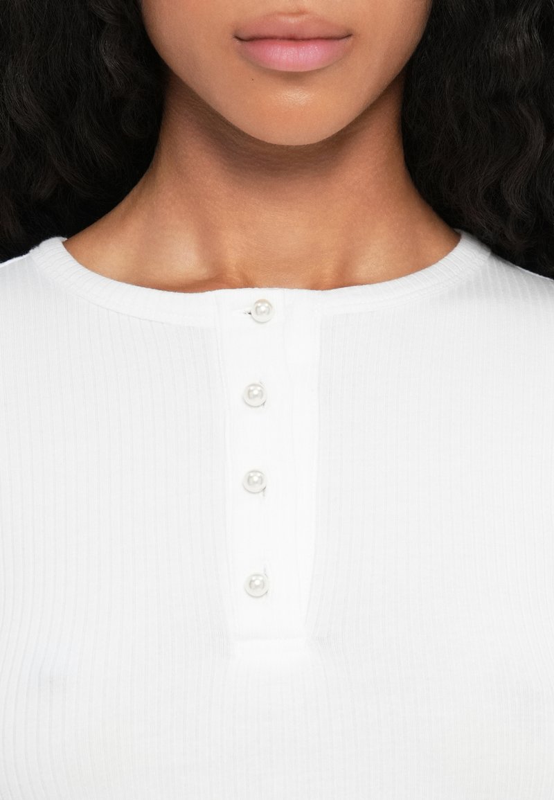 Haut blanc à côtes avec un col rond et une rangée de cinq boutons en perles sur le devant. Texture lisse, coupe ajustée.
