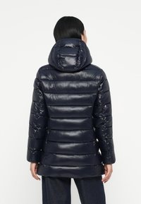 Veste rembourrée bleu marine avec une finition brillante, capuche ajustée et un motif matelassé horizontal. Tissu durable, longueur mi-longue, manches longues et poches latérales.