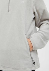 Hellgrauer Fleece-Pullover mit einem halben Reißverschluss, der eine Seitentasche und kontrastierende Reißverschlussdetails aufweist. Weiche Textur und einfaches Design.
