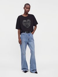 Άτομο που φοράει μαύρο t-shirt με καρδιά και κείμενο και ανοιχτό μπλε φαρδιά τζιν με σκισμένα τελειώματα, που στέκεται μπροστά σε απλό φόντο.