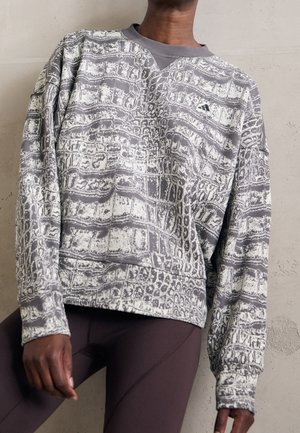 Persoon draagt een grijs-wit patroon sweatshirt met een klein Adidas-logo, gecombineerd met donkere aansluitende leggings, tegen een effen muur als achtergrond.